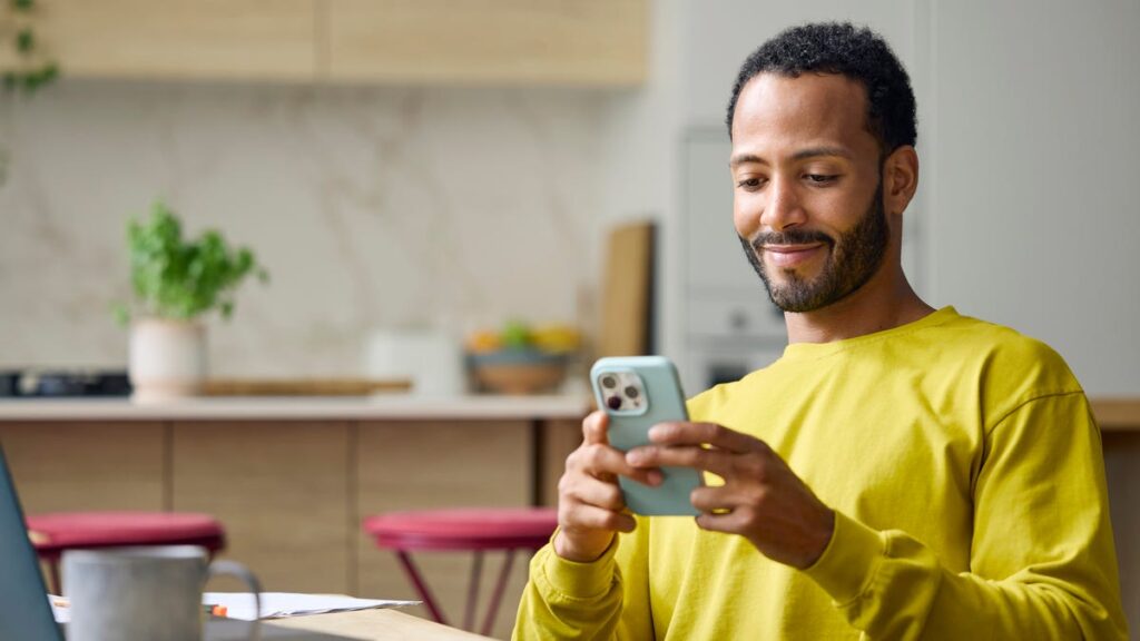 A man in yellow smiles at his phone while working in the kitchen.