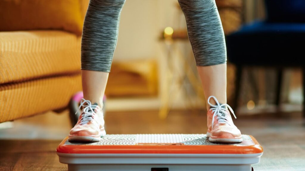 A close-up of a person wearing grey leggings and sneakers standing on a vibration plate in a living room with a mustard yellow couch.
