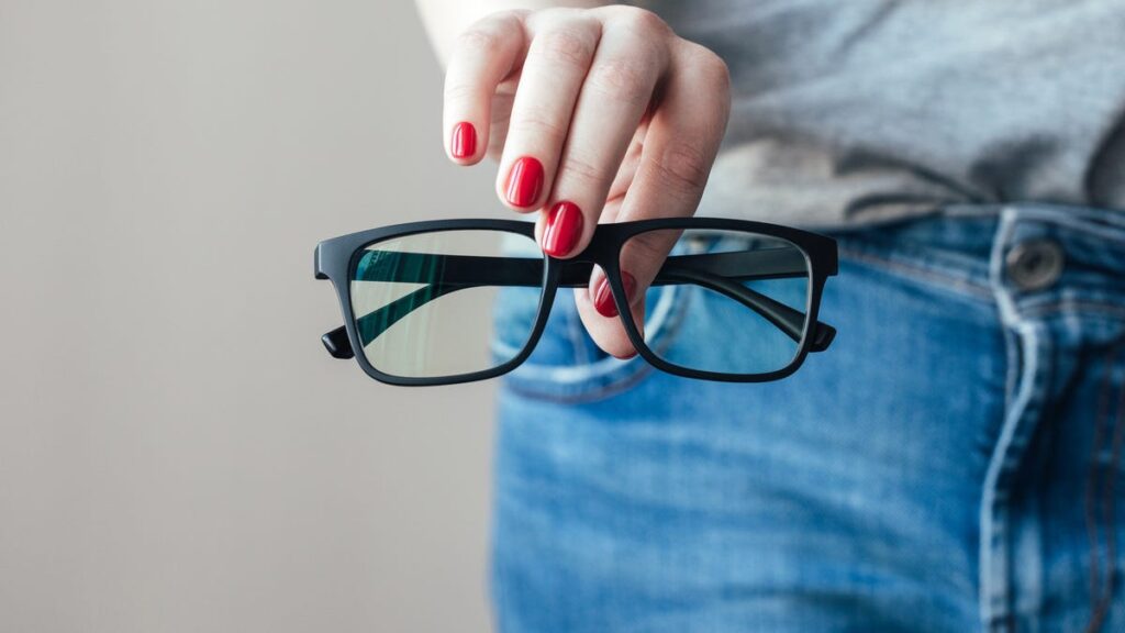 A close-up on a hand with red nail polish holding a pair of black glasses in front of their blue jeans and grey tee.