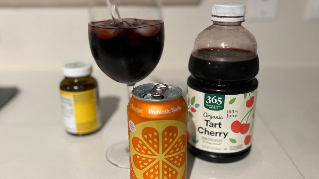 An orange Poppi can sits next to a bottle of tart cherry juice and a wine glass with a dark red liquid inside.