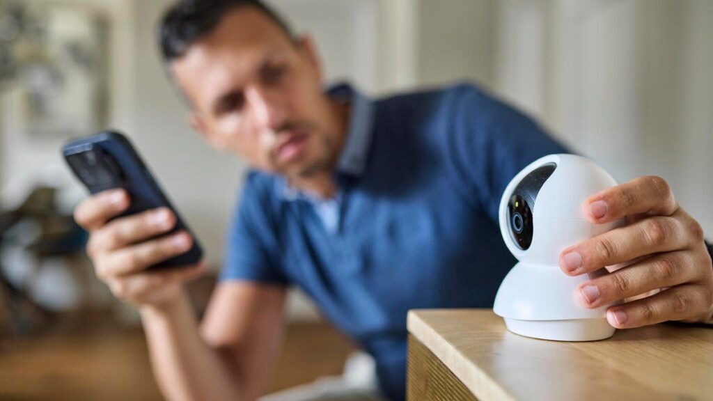 A man holding a phone looks a security camera he is holding on a wooden home table.