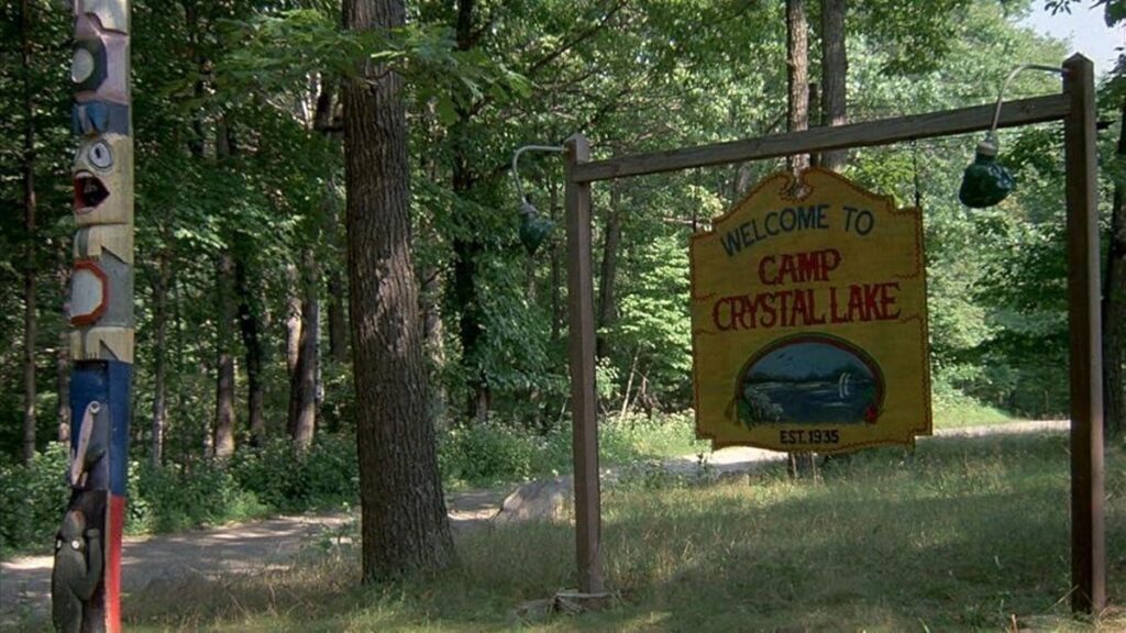 in a screenshot from the movie Friday the 13th, a sign reading Welcome to Camp Crystal Lake hangs on a simple wooden frame next to a native american totem pole