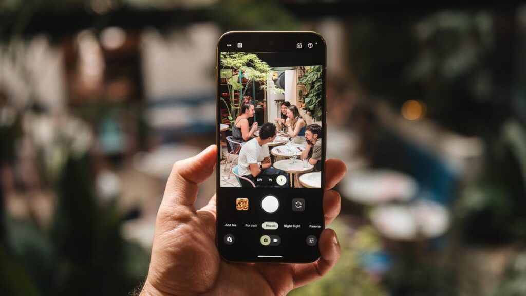 A hand holds a Pixel 10 Pro XL taking a photo of an outdoor dining scene.
