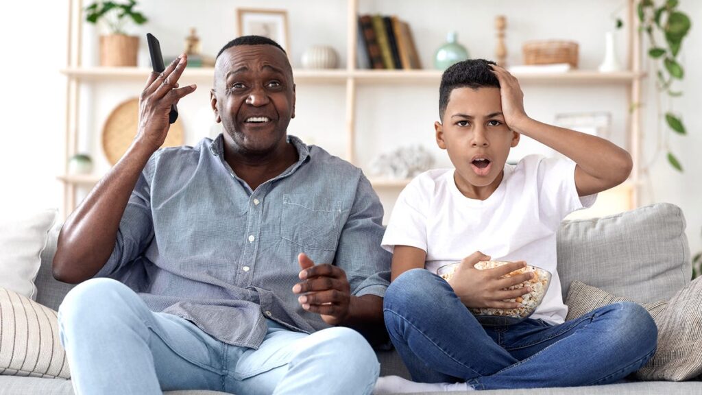 An older man and a boy on a couch looking annoyed and shocked.