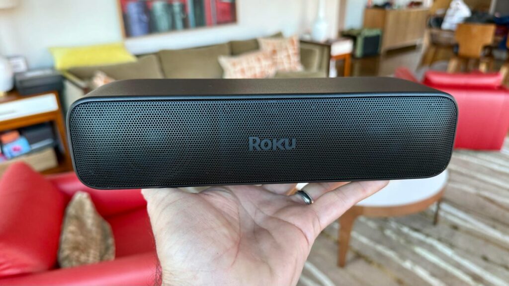 A hand holding a black, oblong, Roku-branded speaker, with a living room visible in the blurred background.