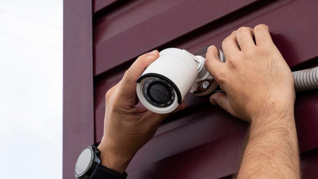 Hands place a security cam on a base on exterior siding.