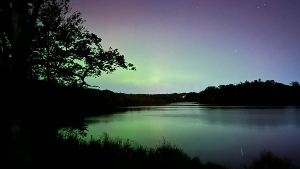 Green aurora reflected in a body of water with trees along the side.