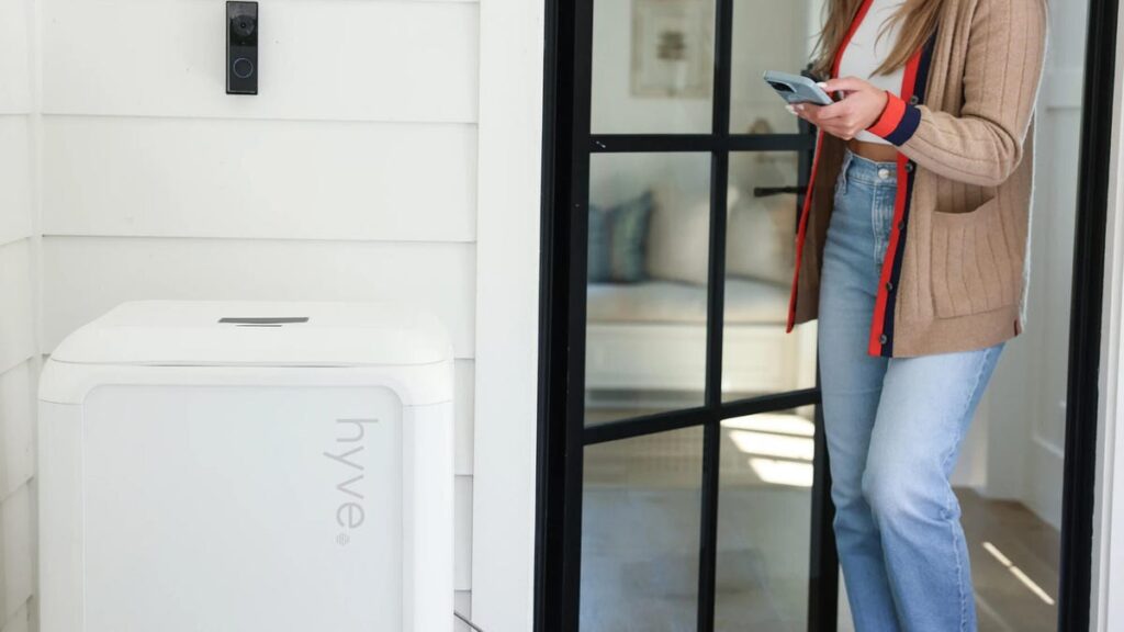 A woman approaches a white Hyve box by a white wall with her phone out.