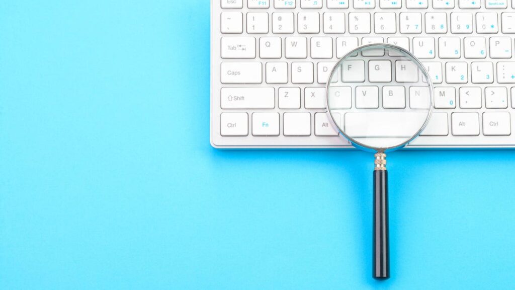 a photo of a keyboard on a blue background, with a magnifying glass on top of the keyboard (gettyimages-1372176504)