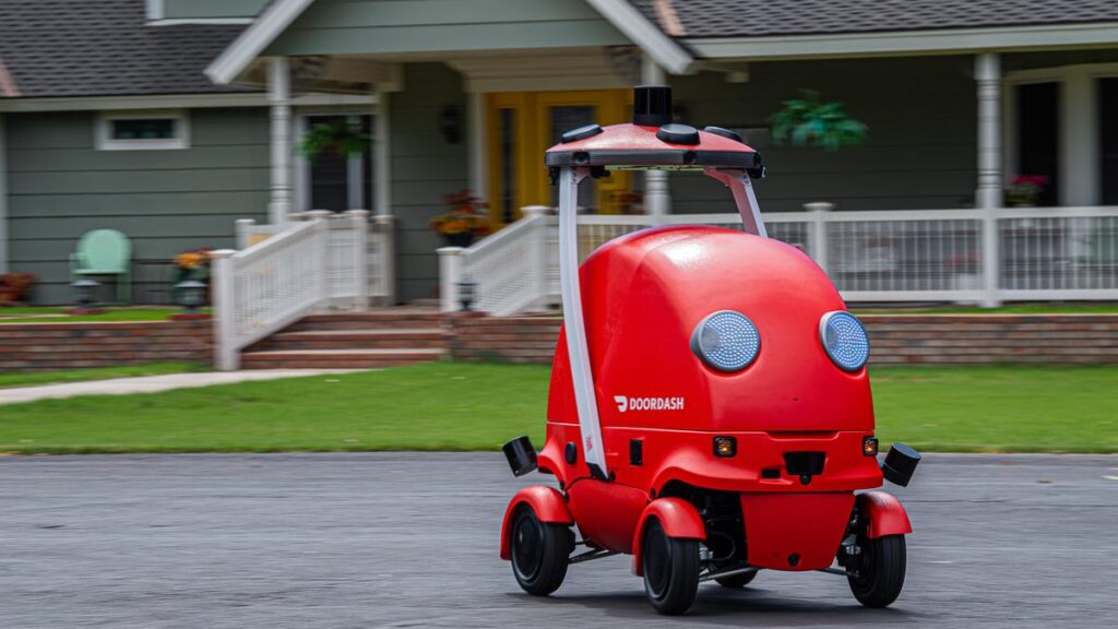 The red DoorDash Dot robot rolls down a street in an idyllic suburban neighborhood.