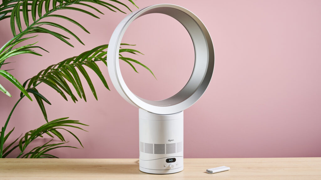 The Dyson Cool CF1 is facing slightly to the right on a table top in front of a leafy green plant and a pink wall. The remote control is on the table to the right of the fan.
