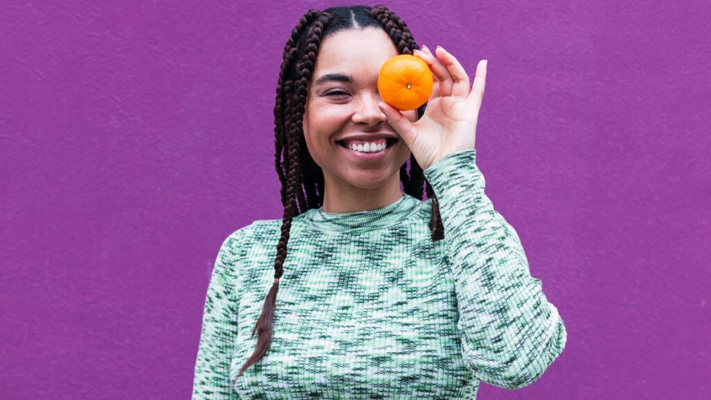 A person with long brown braided hair and a teal patterned shirt holding an orange up in front of their left eye.