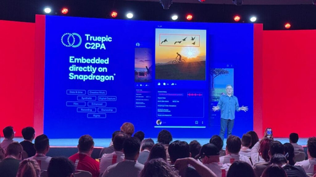 A man stands on stage with a presentation behind him announcing new phone features.