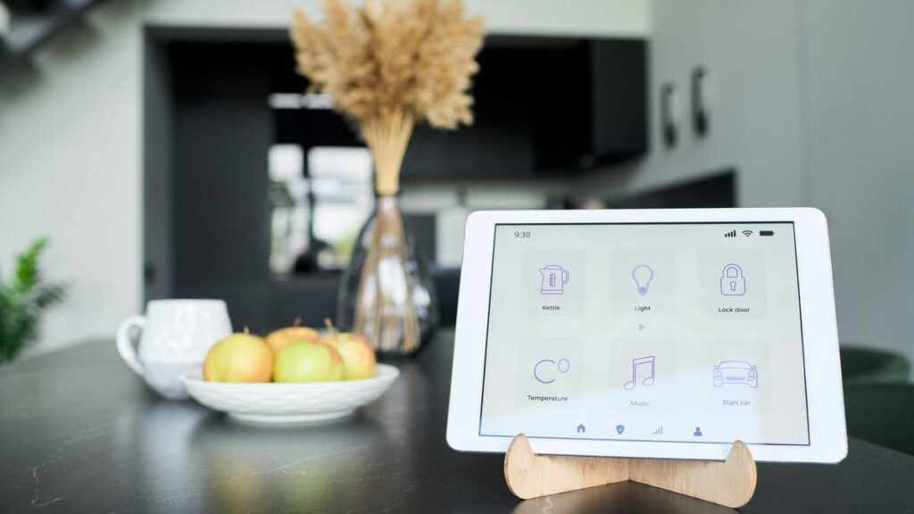 A home smart display on a wood stand on a dark table with a bowl of apples beside it.