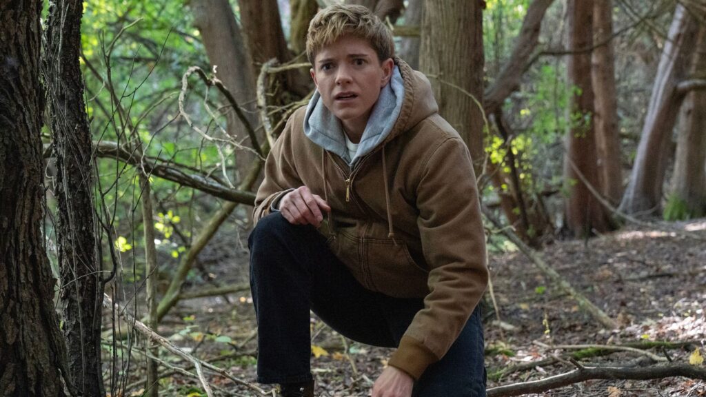 Mae Martin kneels one knee on the ground in the middle of the woods