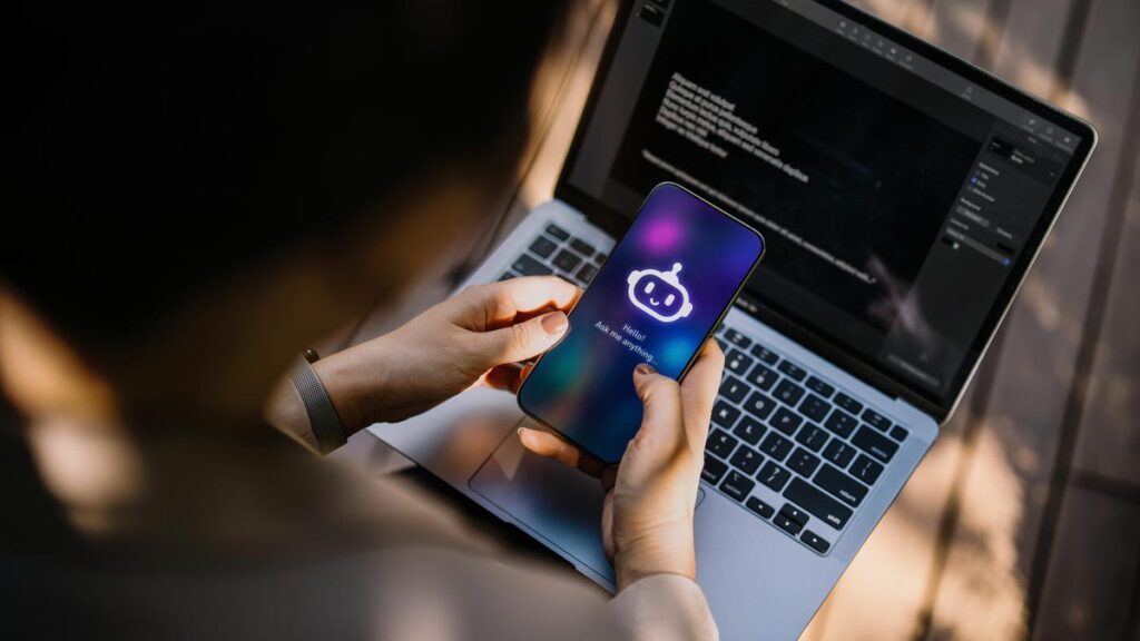 A photo of someone using AI on their phone, with their laptop in the background (gettyimages-2193072141)
