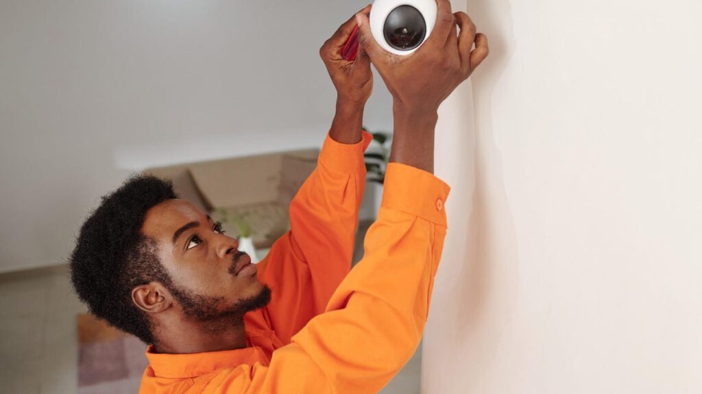 A man in orange reaches up to work on a security camera on a white wall.