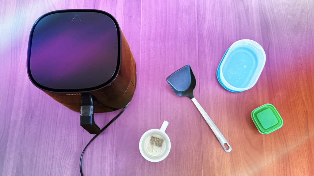 An air fryer, black plastic spatula, mug with a tea bag and plastic containers on a wood surface.