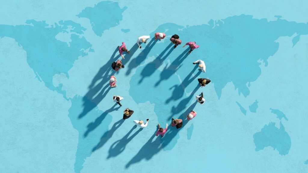 An image of a circle of diverse people standing on a map of the world (gettyimages-1389851402)