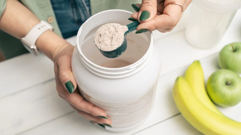 A person with green nails and a smartwatch scooping a white collagen powder out of a container next to a banana and two green apples.