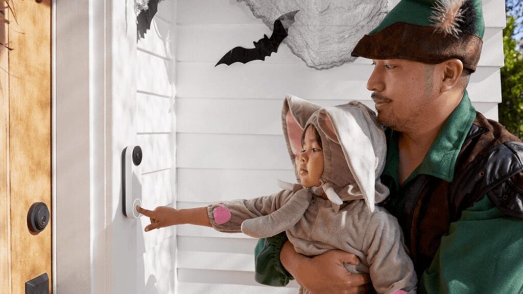 A man holds a child up to a video doorbell in costumers with Halloween decorations.