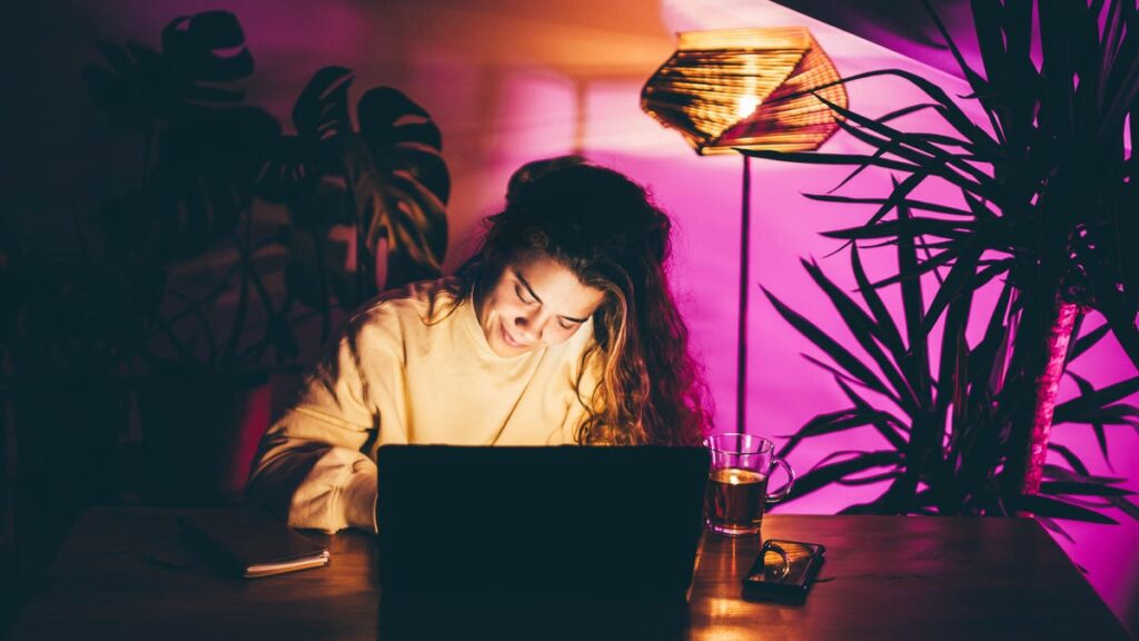 A photo of a woman typing on her laptop, with neon pink lighting behind her (gettyimages-1445395881)