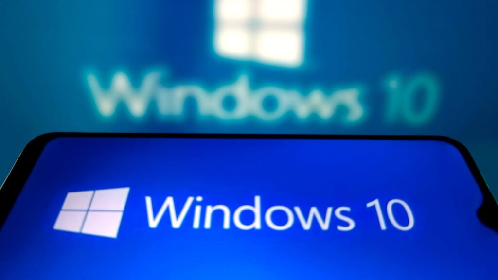 Microsoft Ends Support for Windows 10 Tuesday. Here’s What You Need to Know a Windows 10 logo on a phone with a blue background, held at an angle in front of a blurry Windows 10 logo on a computer screen