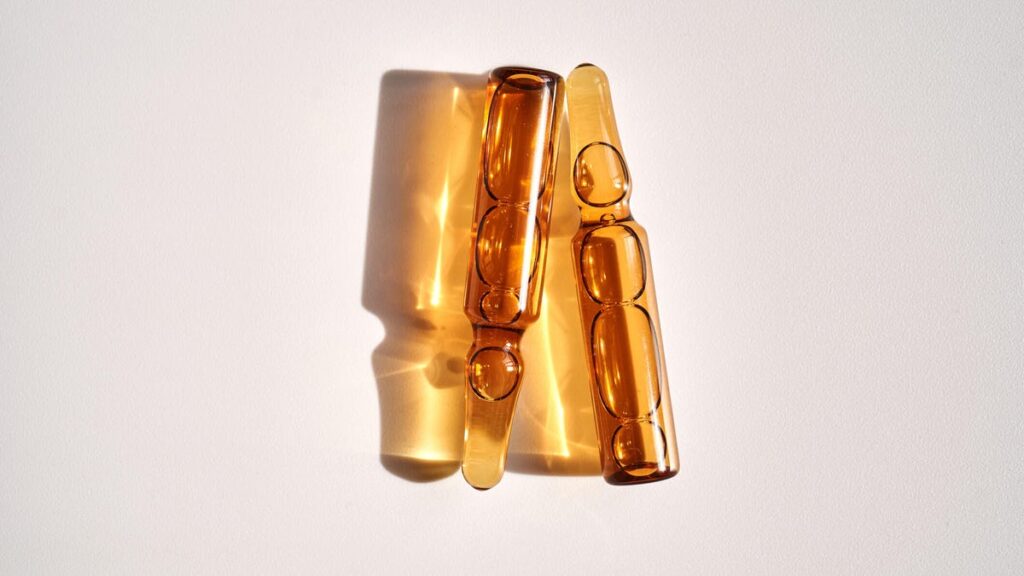 Two brown-colored vitamin D ampoules and their reflections on a white background.