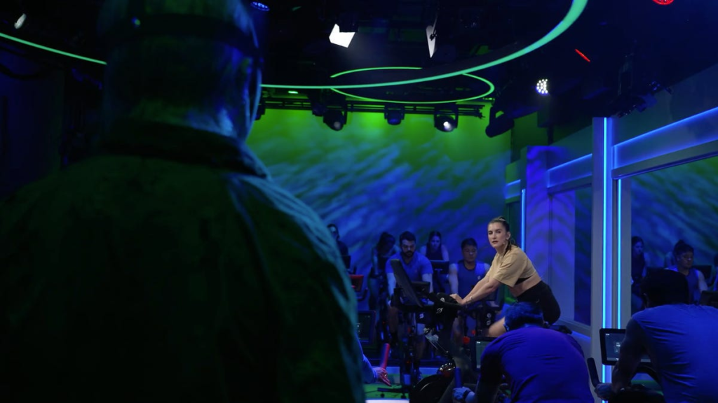 Peloton instructor peers at a masked killer as she cycles away on her exercise bike.