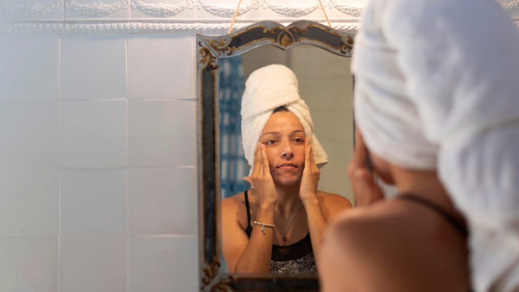 A woman with a towel around her hair looks at herself in the mirror, with her hands on her cheeks