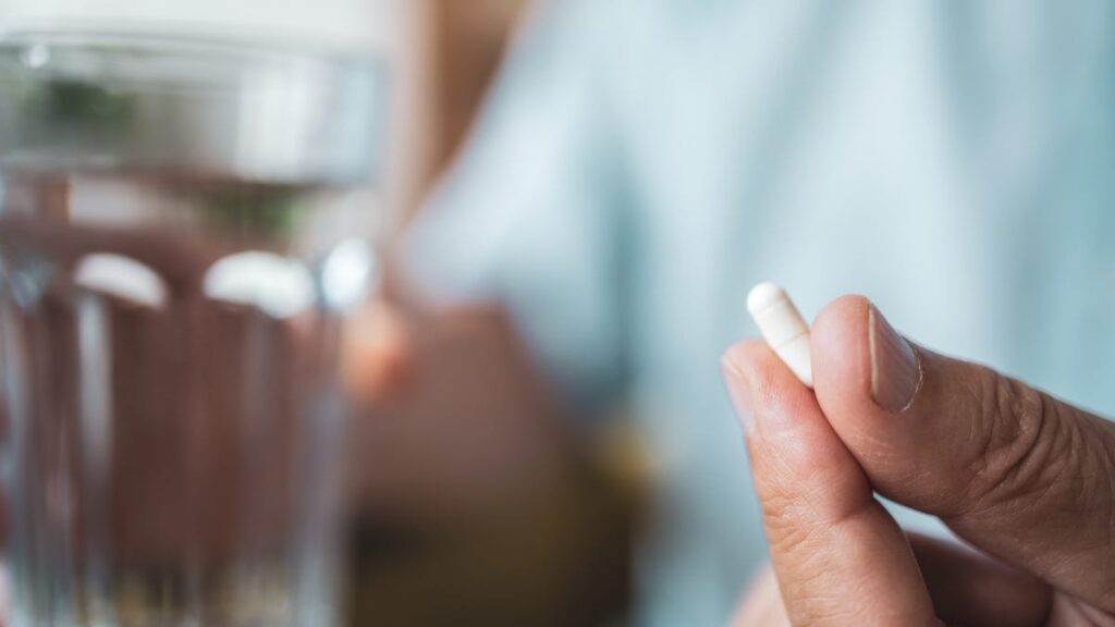 A hand holding a small white capsule with a glass of water in the background.