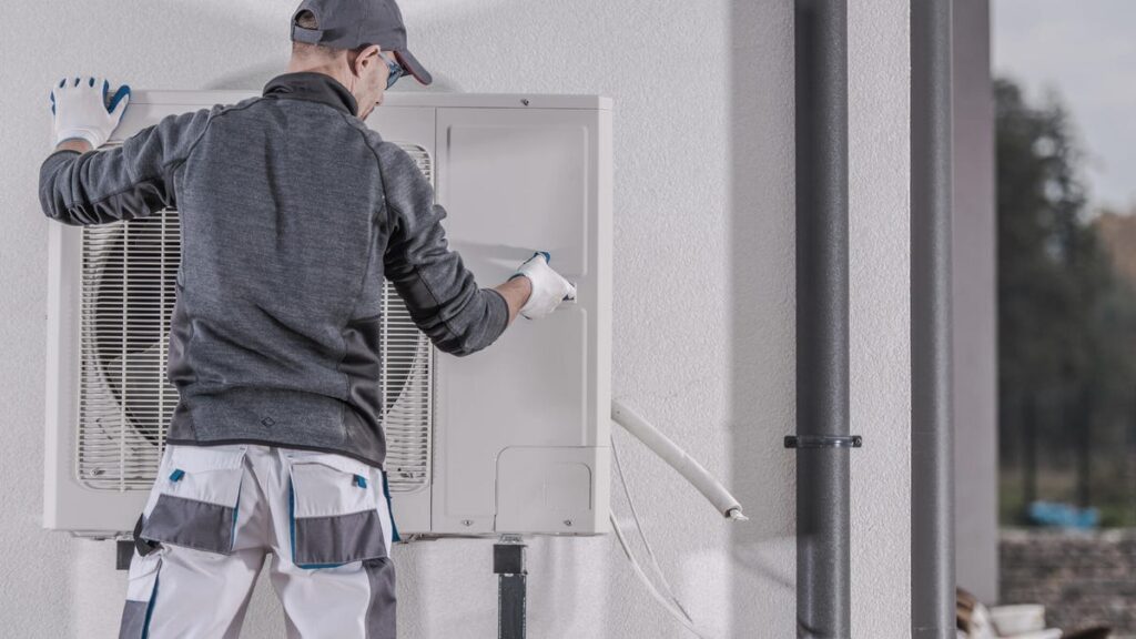 An HVAC installer wearing a gray jacket and hat installs a gray heat pump.